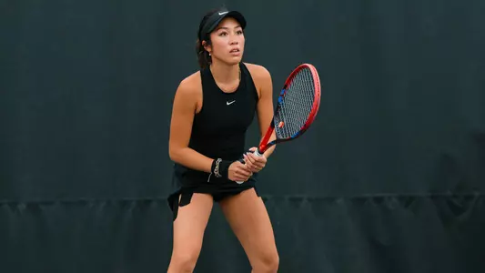 A focused tennis player in a black athletic outfit and visor stands in a ready position on the court, gripping a red and black racket with both hands. She faces forward with an alert expression, knees slightly bent, prepared to receive a shot. The background is a dark, plain court barrier, emphasizing her stance and concentration.