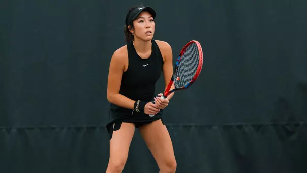 A focused tennis player in a black athletic outfit and visor stands in a ready position on the court, gripping a red and black racket with both hands. She faces forward with an alert expression, knees slightly bent, prepared to receive a shot. The background is a dark, plain court barrier, emphasizing her stance and concentration.