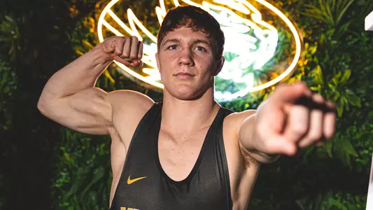 Zan Fugitt in a black singlet poses confidently, flexing one arm while extending the other toward the camera. He stands in front of a glowing neon tiger sign, with green foliage in the background, creating a bold, high-contrast portrait.