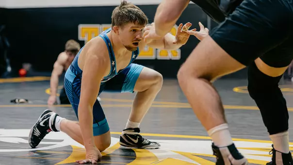 Jarrett Jacques in a blue singlet crouches low on one knee during a match, reaching forward with focus toward an opponent whose legs are visible in the foreground. They are on a wrestling mat inside a gym, with another athlete blurred in the background.