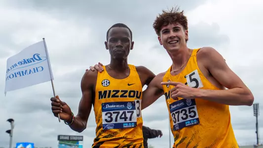 Elijah Limo holds Drake Relays Champion flag after winning 3000m Steeplechase at Drake Relays. Joshua Allison stands next to him and points at him.