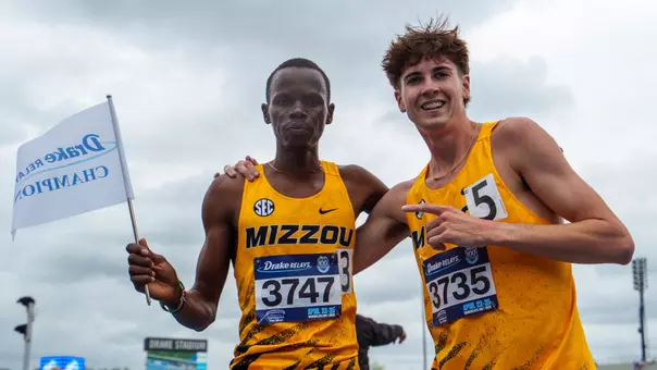 Elijah Limo holds Drake Relays Champion flag after winning 3000m Steeplechase at Drake Relays. Joshua Allison stands next to him and points at him.