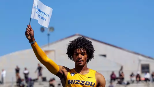 The image depicts Dillon Leacock, wearing a yellow “Mizzou” uniform stands on a podium, raising a white flag that reads “Drake Relays Champion.” A blue bulldog statue covered in signatures sits in front of him, with stadium bleachers and spectators in the background under a bright blue sky.