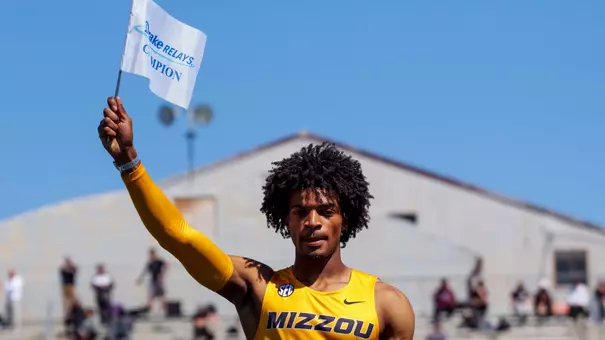 The image depicts Dillon Leacock, wearing a yellow “Mizzou” uniform stands on a podium, raising a white flag that reads “Drake Relays Champion.” A blue bulldog statue covered in signatures sits in front of him, with stadium bleachers and spectators in the background under a bright blue sky.