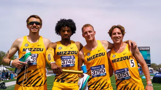 Four male track athletes, Lasse Funck, Dillon Leacock, Finn Russell and Carter Spradling, wearing matching yellow “Mizzou” uniforms stand arm-in-arm on a track field, smiling at the camera. One athlete holds a relay baton, and two hold running shoes. Race bibs are visible on their chests, and a stadium and sky appear in the background.