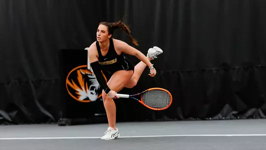 A female tennis player in a black Mizzou uniform lunges forward mid-swing on an indoor court, her body low and balanced as she tracks the ball. She grips a bright orange racket with both hands, her hair pulled back in a ponytail, and wears white tennis shoes. A large tiger logo and dark curtain backdrop fill the background, emphasizing the intensity and focus of the moment.