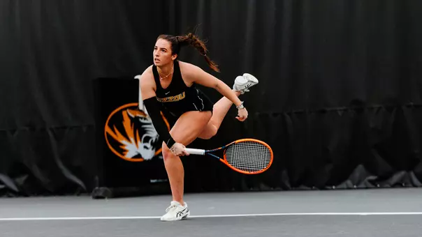 A female tennis player in a black Mizzou uniform lunges forward mid-swing on an indoor court, her body low and balanced as she tracks the ball. She grips a bright orange racket with both hands, her hair pulled back in a ponytail, and wears white tennis shoes. A large tiger logo and dark curtain backdrop fill the background, emphasizing the intensity and focus of the moment.