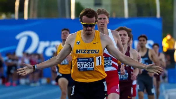 Lasse Funck, a male track runner in a yellow “Mizzou” uniform crosses the finish line with arms outstretched, wearing sunglasses and a race bib, as several competitors sprint closely behind him on a blue track.