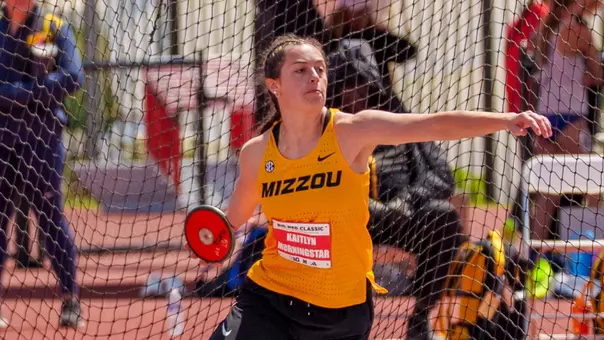 A Mizzou track and field athlete throws a discus inside a netted throwing cage. She wears a yellow “Mizzou” singlet and black pants, with her arm extended mid-throw and her body rotating. A name bib reading “Kaitlyn Morningstar” is visible on her chest, and other athletes and equipment appear blurred in the background.