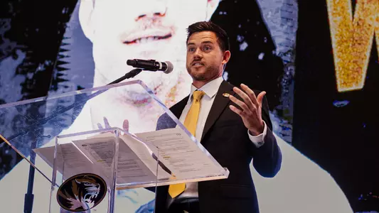 Robin Goodman in a black suit and gold tie speaks at a clear podium with a Mizzou Tigers logo, gesturing with one hand while standing in front of a large screen displaying a close-up portrait behind him. A microphone is positioned at the podium during the indoor event.