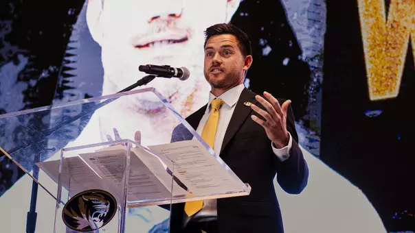 Robin Goodman in a black suit and gold tie speaks at a clear podium with a Mizzou Tigers logo, gesturing with one hand while standing in front of a large screen displaying a close-up portrait behind him. A microphone is positioned at the podium during the indoor event.