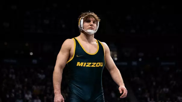 Luke Geog wearing a black and gold “MIZZOU” singlet and white headgear stands under arena lighting, looking focused and determined. He is centered in the frame with a strong, upright posture, while a dark, blurred crowd fills the background, creating a dramatic, spotlight effect around him.