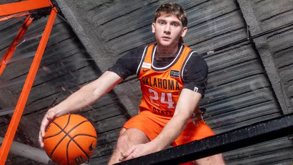Oklahoma State Cowboy Basketball player Ryan Crotty (#24) poses for a promotional photo beneath a glass backboard, holding a Nike basketball with one hand while looking directly into the camera. He wears the team's orange Oklahoma State jersey with the Big 12 conference logo visible on the chest. The photo is taken from a low angle looking up through the backboard, with orange support structures framing the shot.