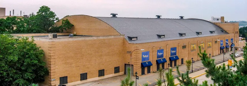 The exterior of Fitzgerald Field House flanked by a nearly empty parking lot and absolutely no street traffic. It is a gray, cloudless sky-- the plant life would suggest spring or summer. The building is a long, beige brick structure with a barrel vault roof clad in dark gray raised ridge material. Royal blue awnings shelter the many exterior doorways.  It is a somber scene and as if the old building knows  its days are numbered.