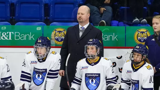 Assistant Coach Brent Hill stands on the bench behind three women's hockey players during a game.