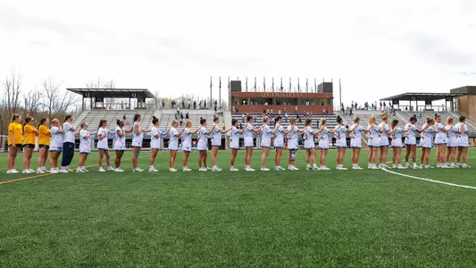 The women's lacrosse team lines up for the national anthem with the Quinnipiac stands in the background before a home game.