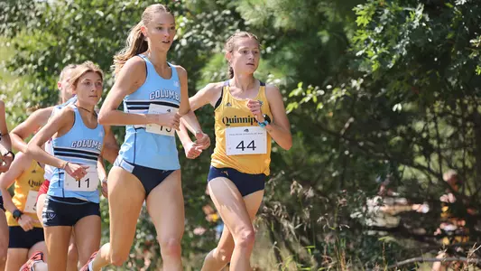 Liv DiStefano, in her gold uniform, runs alongside a Columbia runner in their light blue and white uniform.