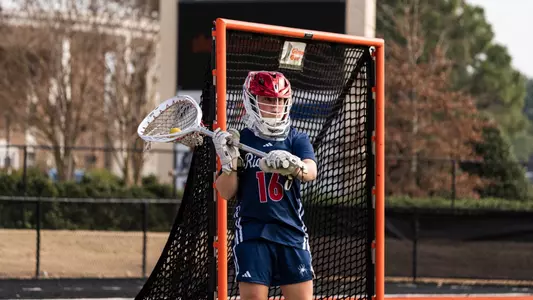 Nicole Burney with the ball in her goalie stick behind her shoulder as she gets ready to pass the ball, the cage is behind her