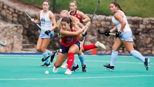 Number 30 Valen Luna Paratore, wearing a red uniform top and blue skirt, winds up to take a shot for Spider Field Hockey at Crenshaw Field