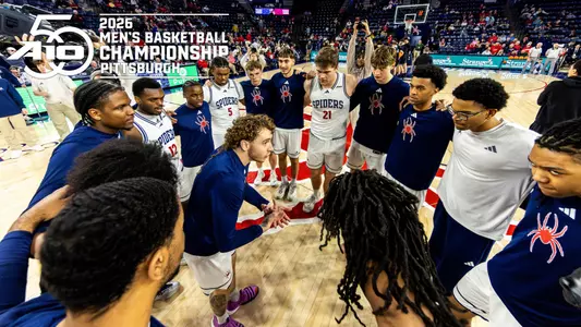 The men's basketball team huddles with the A-10 championship graphic in the upper left