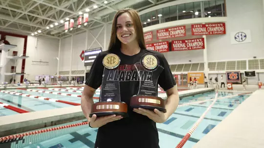 Rhyan White standing at the edge of the pool holding SEC Championship trophies
