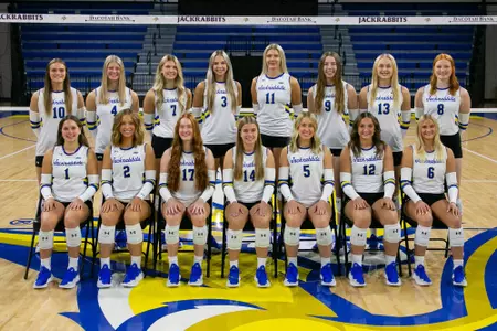 15 volleyball players in white jerseys, facing the camera, posed for a team photo