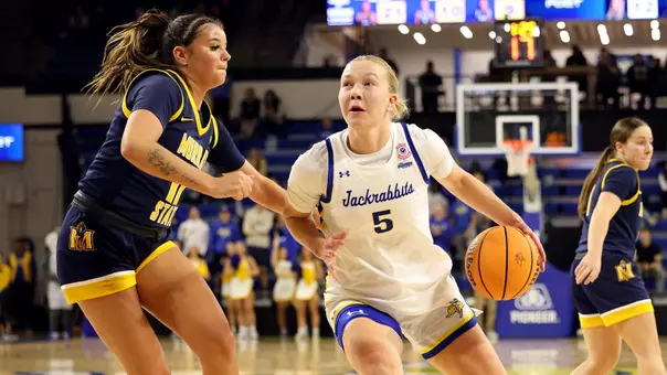 A women's basketball player, wearing a white uniform with "Jackrabbits" written in script, drives to the basket against a defender wearing a navy uniform