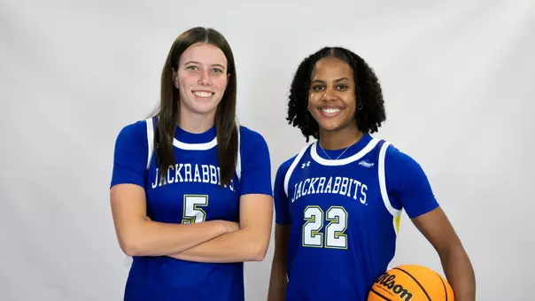 Two women's basketball players wearing blue uniforms pose together in front of a white backdrop