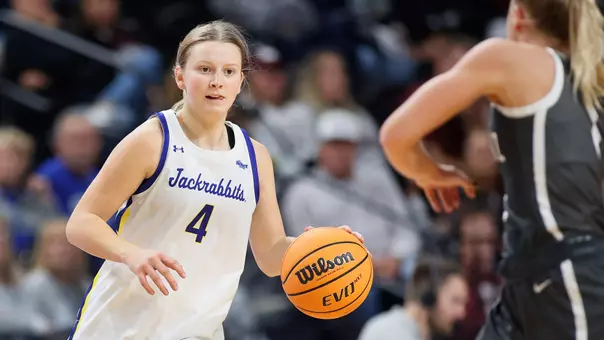 A woman in a white basketball uniform dribbles while an opposing player in a gray uniform defends