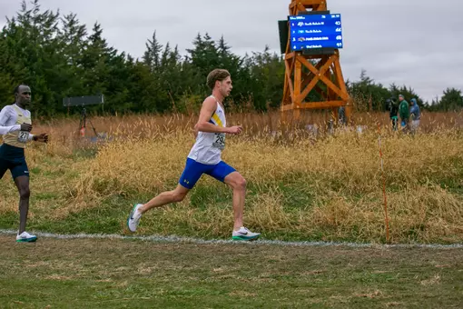 A man runs in front of a windmill and grassy field at a cross country meet
