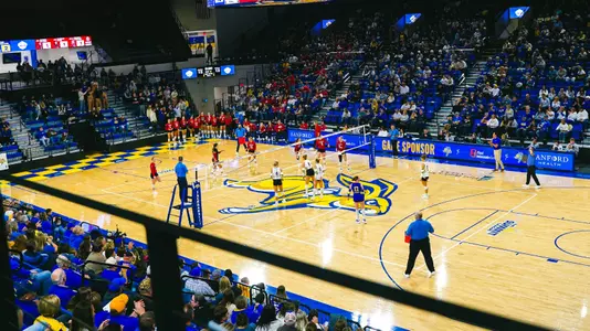 Wide-angle shot of First Bank & Trust Arena during a volleyball match. One side of the net has a team wearing blue uniforms, the other side has a team in red uniforms.