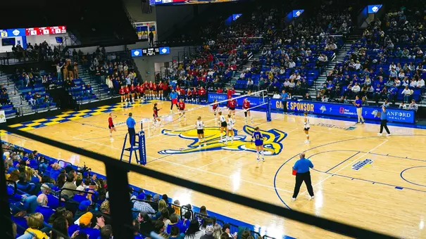 Wide-angle shot of First Bank & Trust Arena during a volleyball match. One side of the net has a team wearing blue uniforms, the other side has a team in red uniforms.