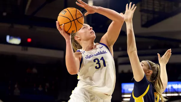 A women's basketball player in a white uniform goes up for a contested shot against a player in a navy uniform