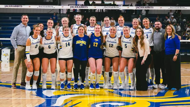 The South Dakota State volleyball team and its staff members pose for a team photo in front of a volleyball net