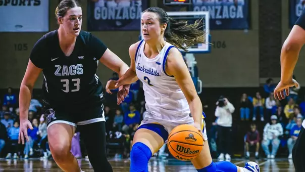 A women’s basketball player in a white jersey dribbles a basketball as she is guarded by an opposing player in a black jersey.