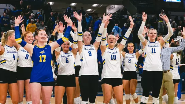 Women's volleyball players in white and blue uniforms wave to the crowd in a sports arena