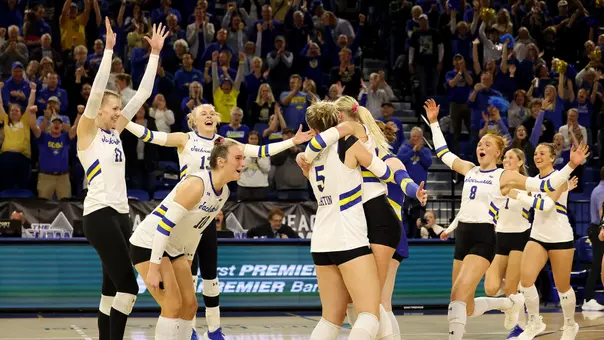 A women's volleyball team in white uniforms celebrates winning a match