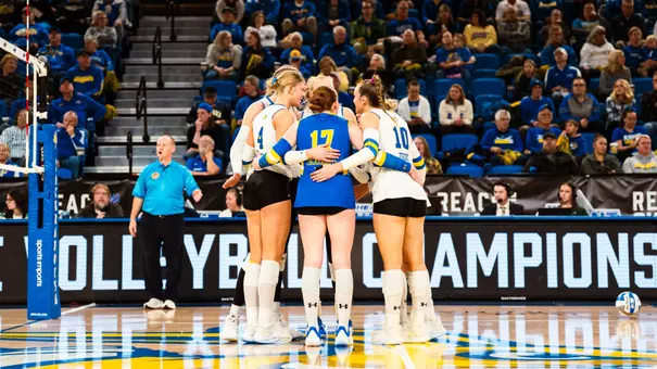 Women's volleyball players in white jerseys huddle on the court with a crowd watching in the stands behind the huddle