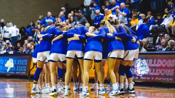 A women's basketball team gathered in a huddle in front of fans in a basketball arena