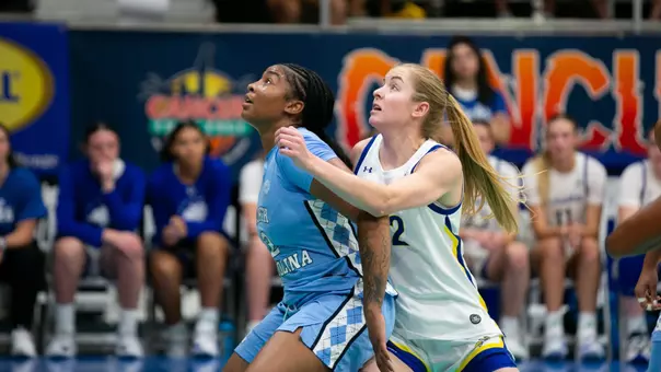 A women's basketball player in a light blue uniform and a player in a white uniform compete for a rebound