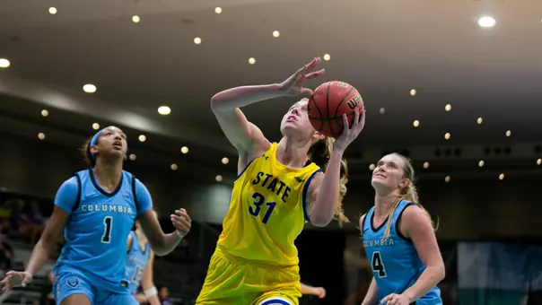 A women's basketball player in a yellow uniform goes up for a shot with two nearby defenders in light blue uniforms