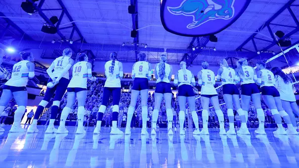 15 volleyball players in white uniforms lineup for the national anthem in a dark arena with blue lighting