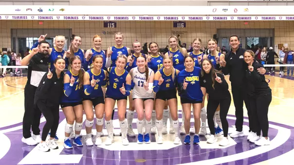 The South Dakota State volleyball team, in blue uniforms, poses at a volleyball net after a match, holding up number-one signs