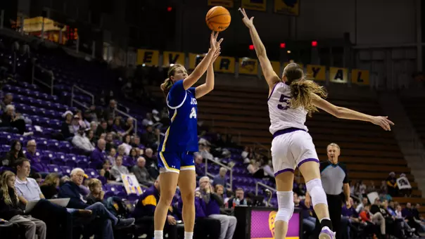 A women's basketball player in a blue uniform shoots a 3-pointer over a player in a white uniform