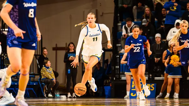 A women's basketball player dribbles up the court with two opposing players also in frame