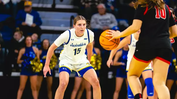 A women's basketball player in a white uniform, facing the camera, defends as a player in a black uniform dribbles
