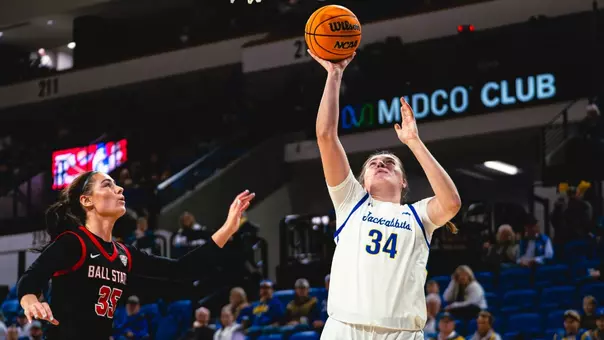 Basketball player in a white jersey with number 34 jumps for a shot during a game. A player in a black uniform is nearby defending