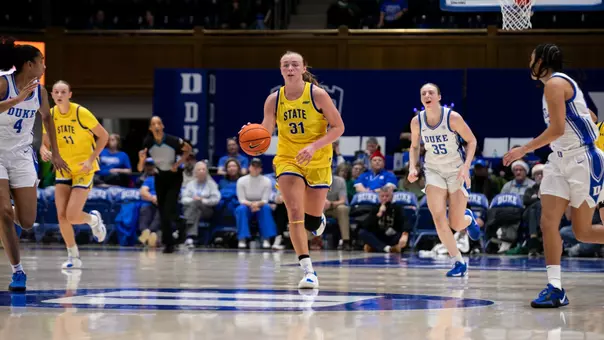 A women's basketball player, Brooklyn Meyer, in a yellow jersey dribbles the basketball up the court, surrounded by Duke players in white uniforms during a game.