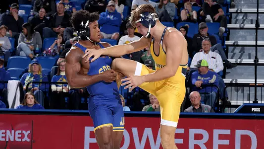 A wrestler in a blue singlet, Marcus Espinoza-Owens, grabs the leg of another wrestler in a yellow singlet, putting it under his arm.