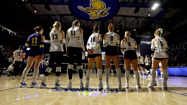 Seven volleyball players in white uniforms stand on the court facing the net, away from the camera